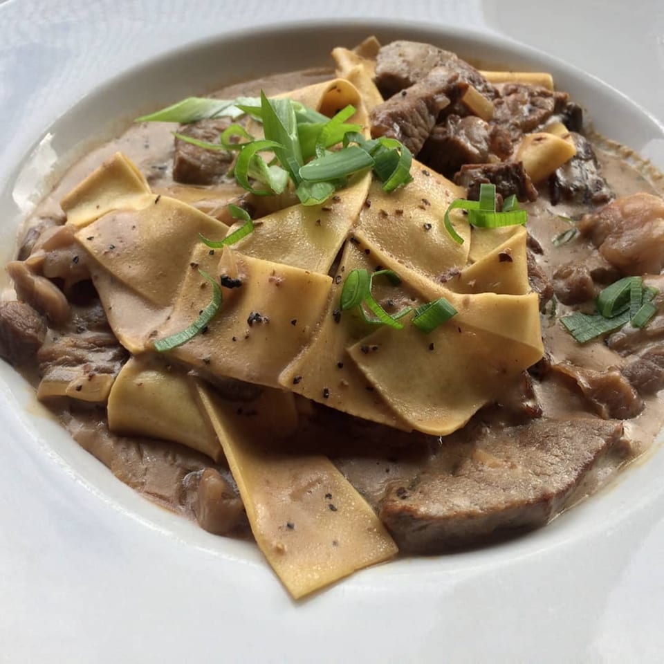 Beef stroganoff with pappardelle pasta, mushrooms, and green onions in a white bowl.