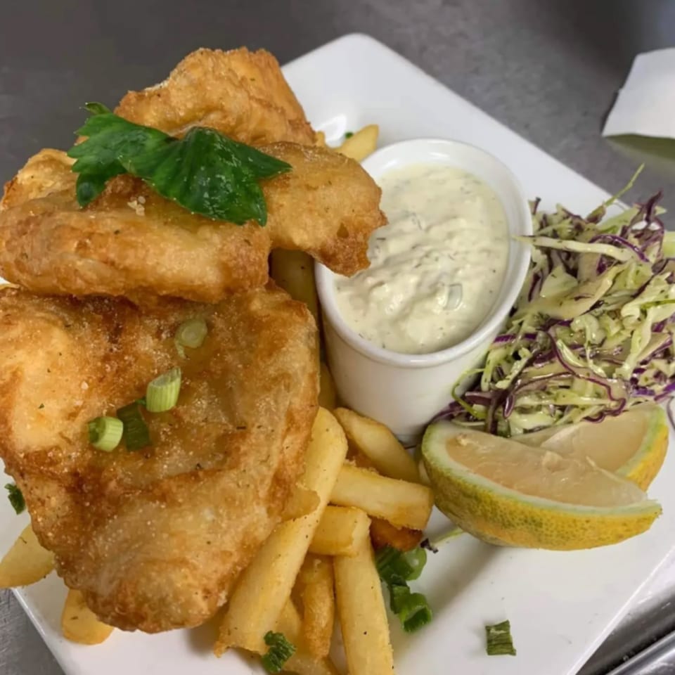 Golden-brown fish and chips with tartar sauce, coleslaw, and a lemon wedge on a white plate.